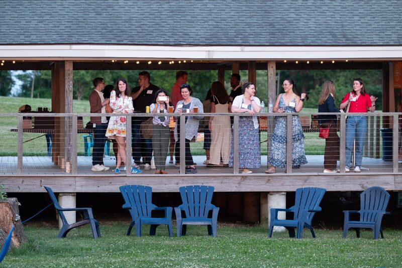 The image shows a group of people gathered on a wooden deck. Some are standing, while others are leaning against a railing. There are blue chairs in front of the deck. The scene appears to be outdoors, possibly at a social gathering or event. The lighting suggests it might be dusk or early evening.